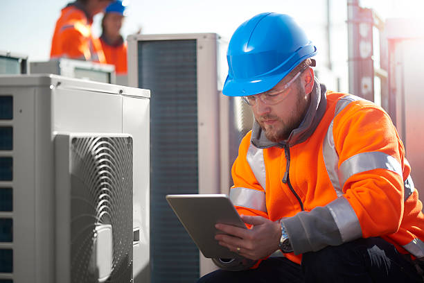 A man wearing an orange vest and hard hat is focused on using a tablet in a work environment.